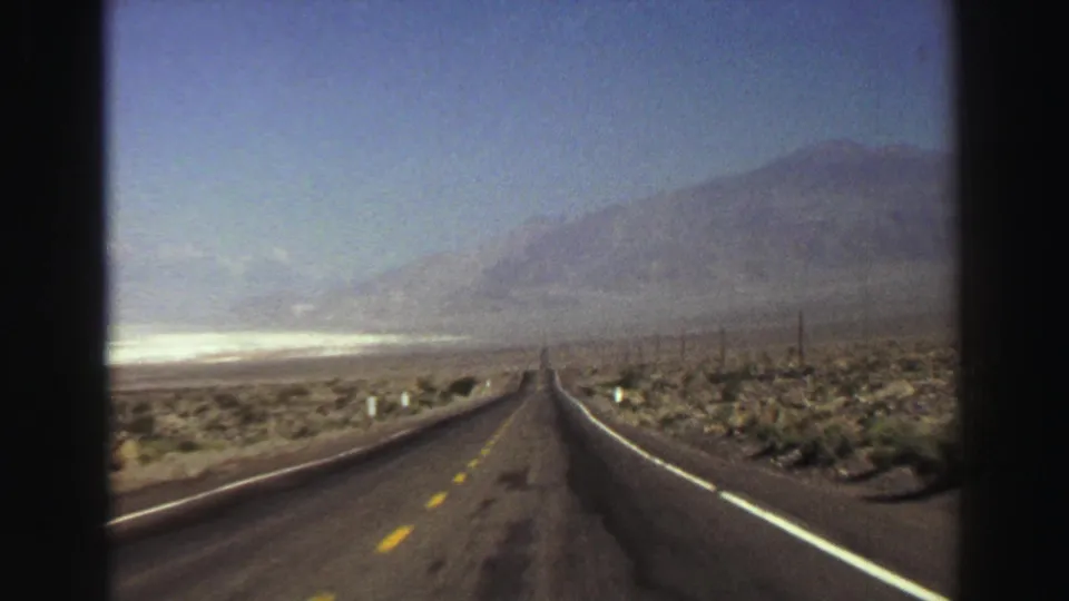 Final 1969 8mm road-trip frame showing a quiet highway through arid scrubland with mountains beyond and no visible people or vehicles nearby.