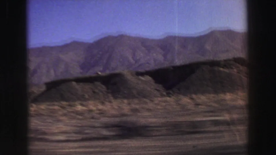 Moving vehicle view of arid roadside hills and distant mountains, with 8mm grain and dark scan borders visible.