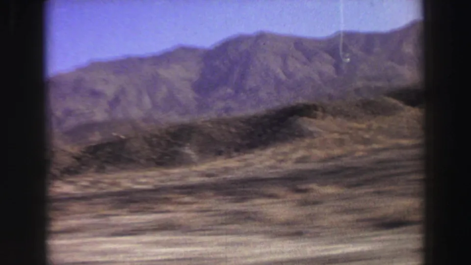 End frame of a moving 8mm road view, with dry terrain in the foreground and a mountain ridge under clear sky.