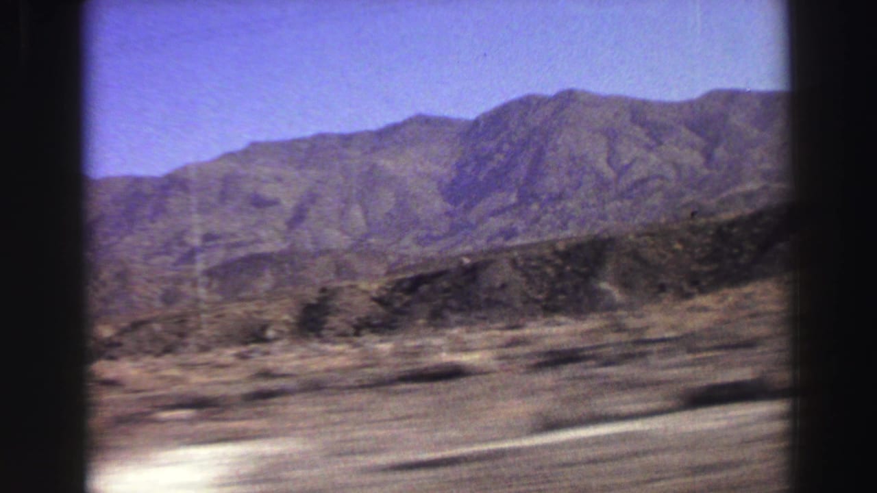 Scrub-covered open land and rocky mountains seen from a moving vehicle.