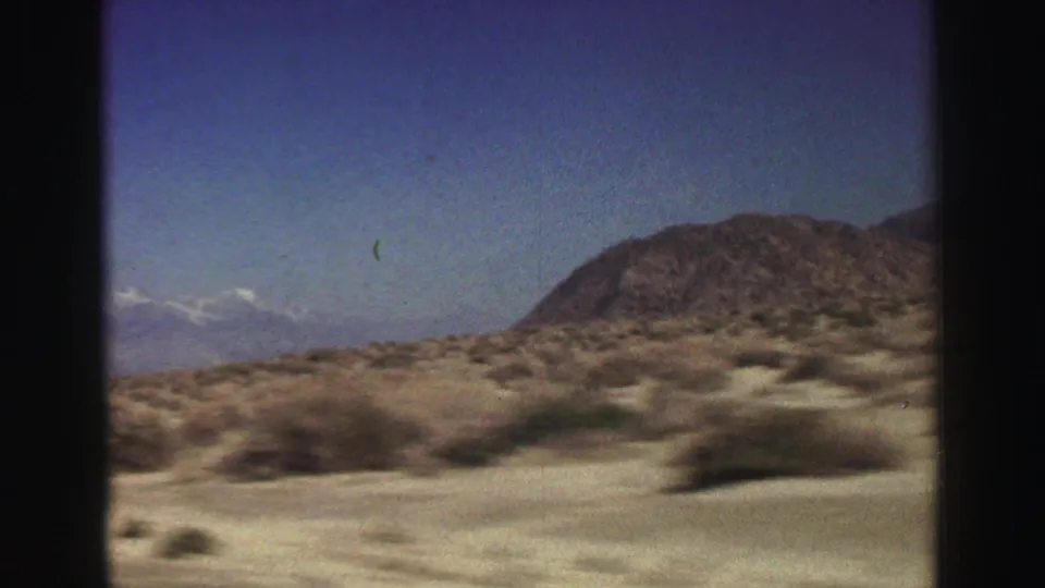 1969 Lake Tahoe-area home-movie frame of dry scrubland sliding past a rocky hill, with distant mountains at left.