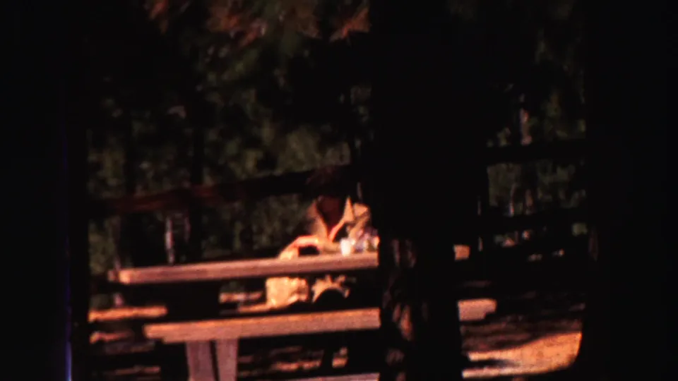 In the 1969 home-movie footage, one person appears seated at a picnic table under trees in a shaded outdoor setting.