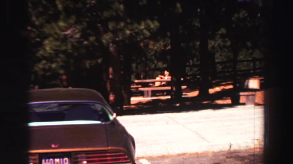 A late-1960s Lake Tahoe park view shows a parked car, California plate, picnic tables, pine trees, and sunlit pavement.