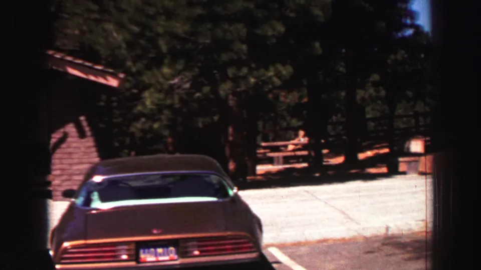 The camera frames the rear of a parked car near a small park structure, picnic tables, pines, and a bright paved area in Lake Tahoe.