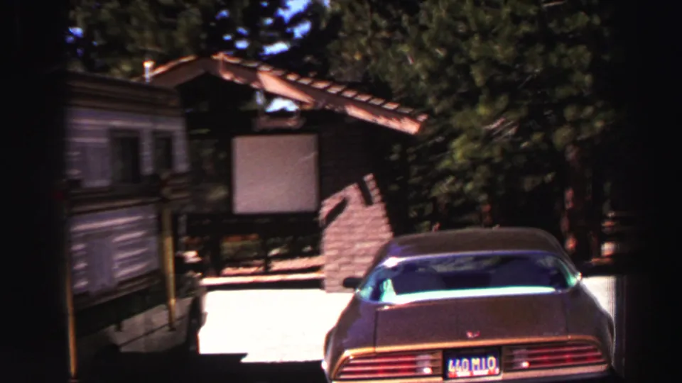 A Lake Tahoe park frame shows a parked car, small building, blank signboard, stone wall, pine trees, and bright pavement.