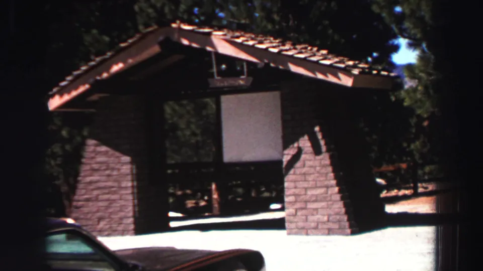 A rustic Lake Tahoe campsite shelter appears behind a partly visible brown car, framed by dark pine woods and bright sun.