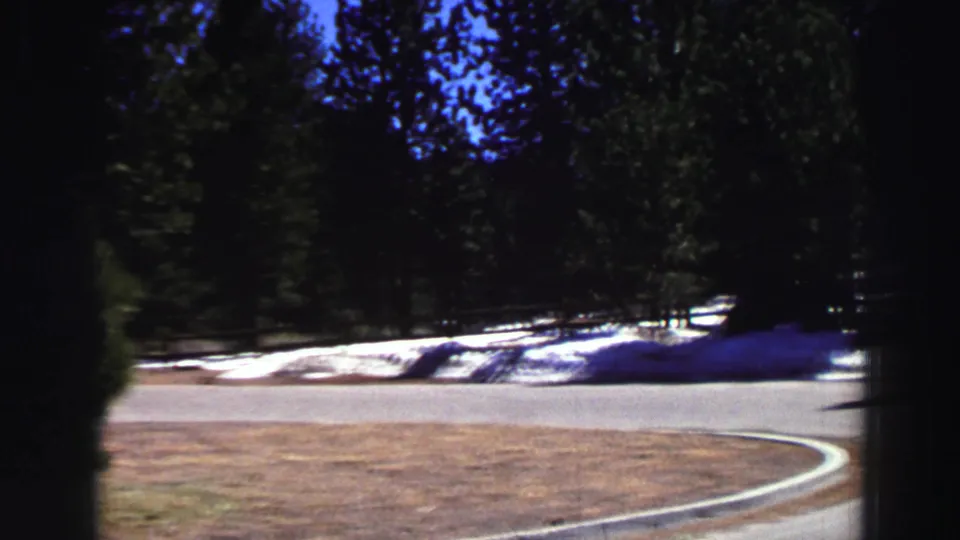 Lake Tahoe roadside view with a bright road curve, evergreen forest, and remaining snow from 1969 home-movie footage.