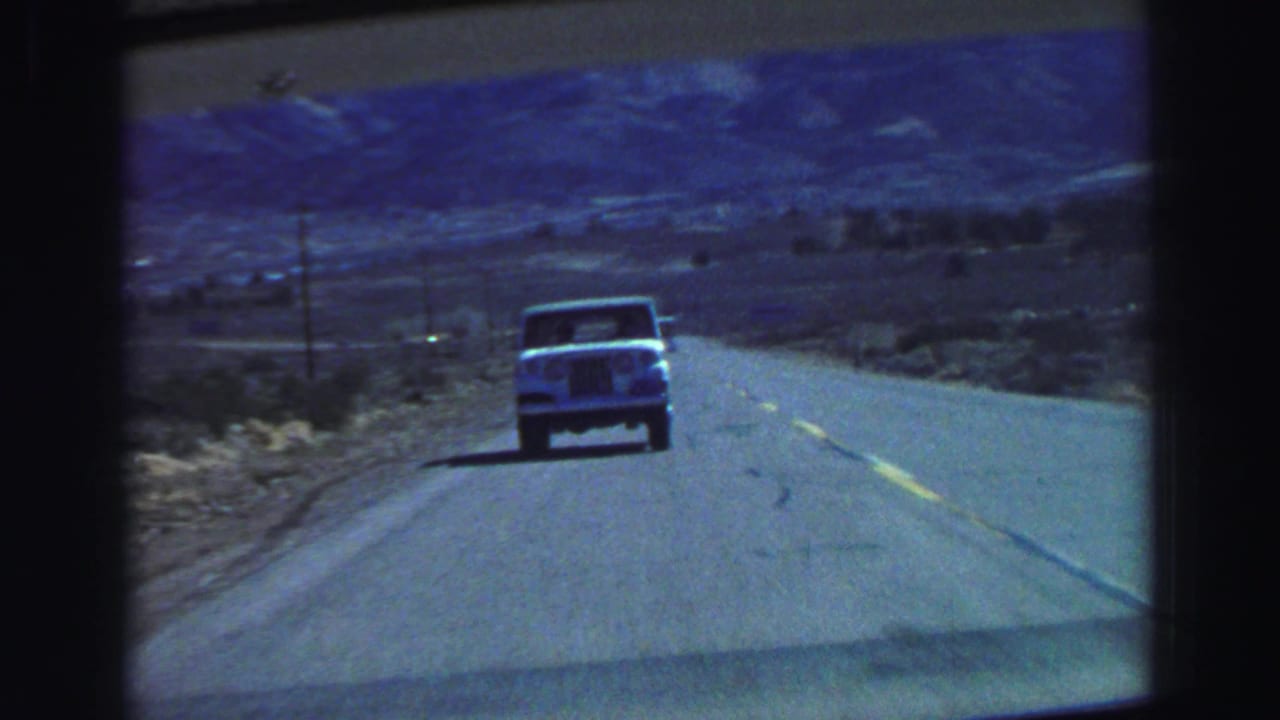 Light-colored truck driving ahead on a two-lane highway with mountains in the distance.