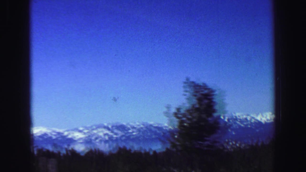 1969 Lake Tahoe travel footage shows snow-capped mountains beyond pine forest, framed by dark edges from a moving viewpoint.