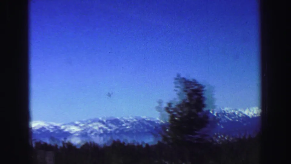 1969 Lake Tahoe travel footage shows snow-capped mountains beyond pine forest, framed by dark edges from a moving viewpoint.