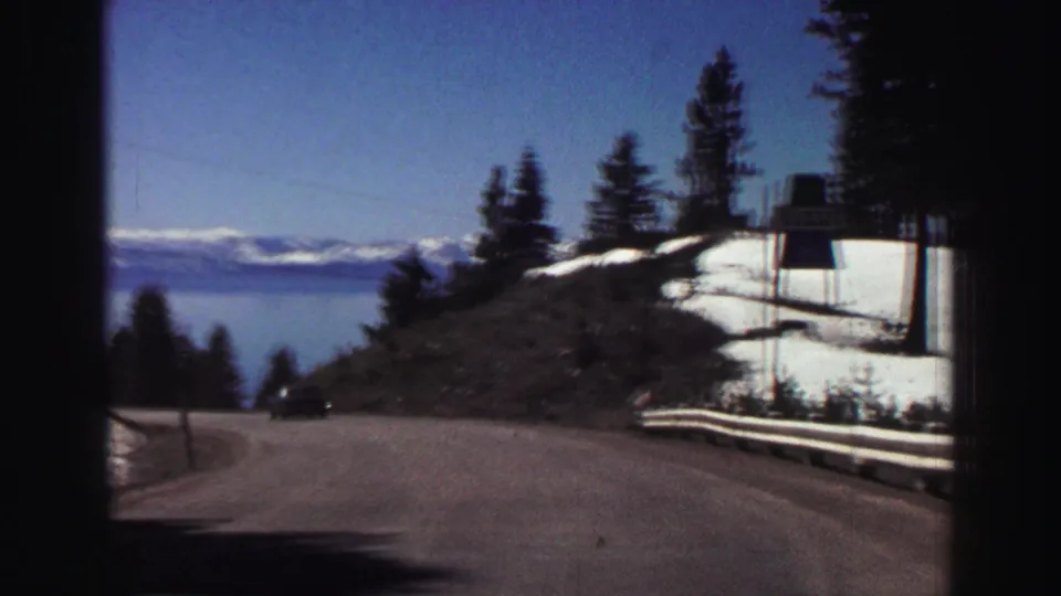1969 Lake Tahoe road view shows a curve above the lake, roadside snow, pines, and distant snow-capped mountains.