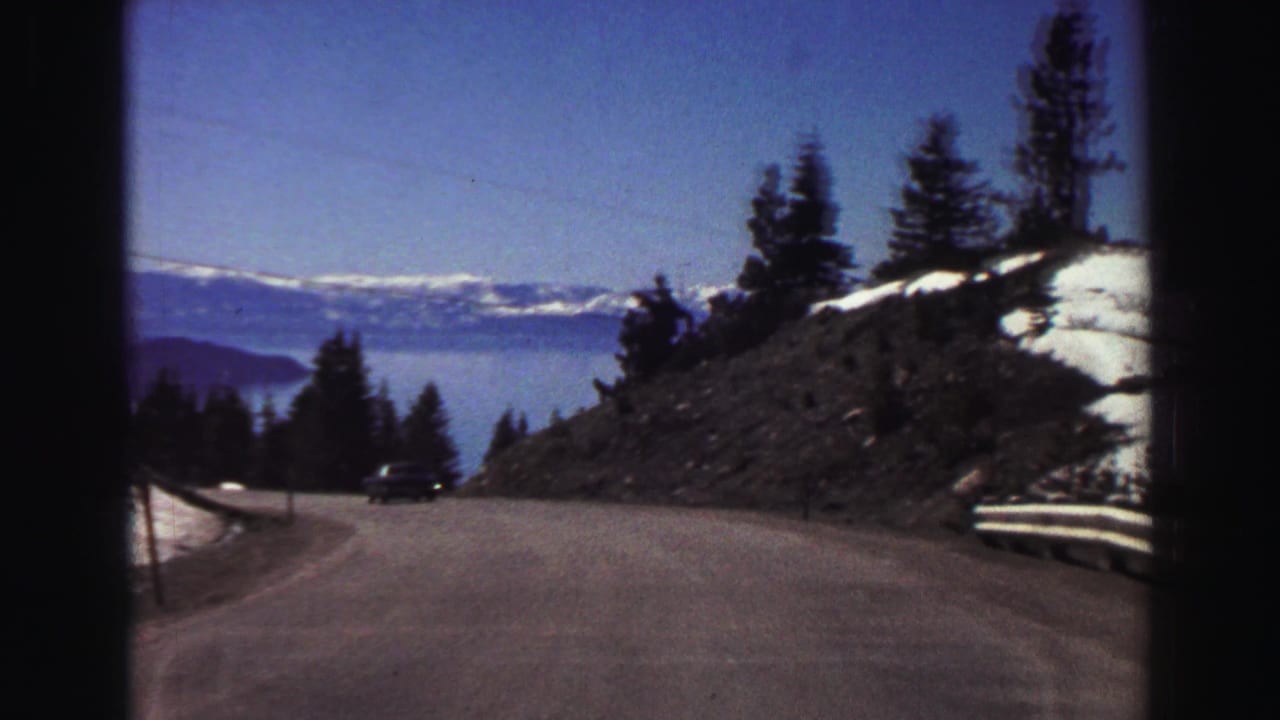 Curving mountain road with snow patches, evergreen trees, distant lake, and mountains.