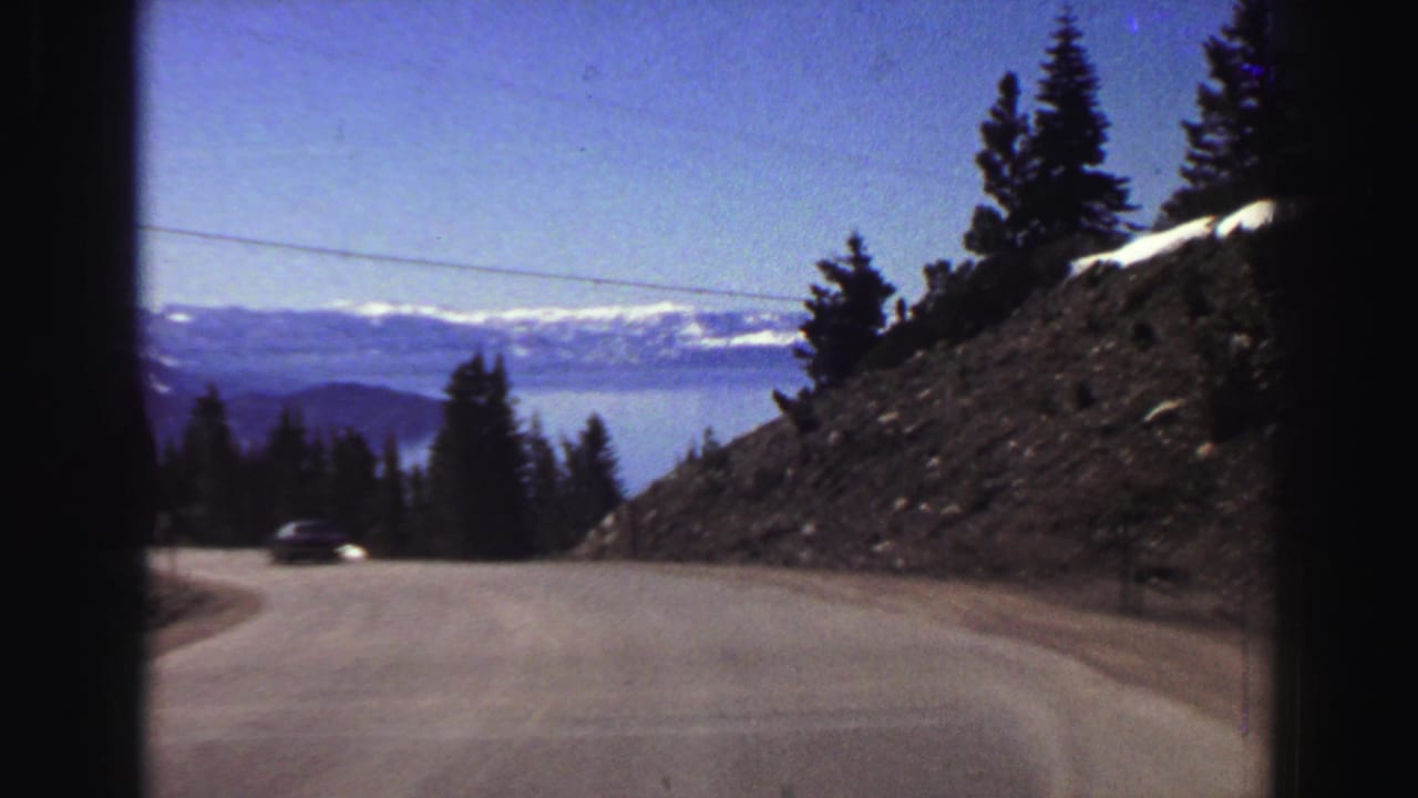A car travels around a paved curve on a Lake Tahoe-area mountain road, with evergreen trees, exposed slope, distant water, and snow-topped mountains visible.