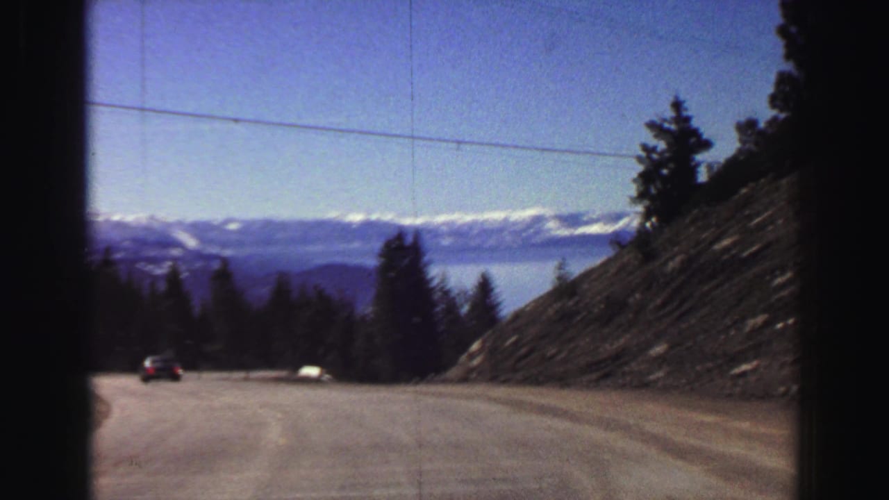 A descending paved mountain road appears to lead toward Lake Tahoe, framed by evergreens, a rocky cut slope, distant water, and snow-capped ridges.