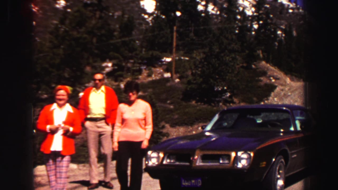 Three adults in late-1960s-style clothing stand beside a dark car in the Lake Tahoe-area roadside setting, with evergreens, snow patches, and gravel visible behind them.