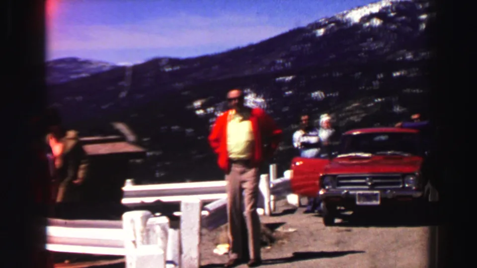 At the Lake Tahoe overlook, a man in a red jacket stands near a red vintage car as other adults remain by the roadside rail and mountain view.