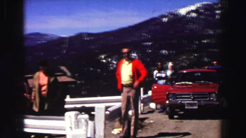 The 1969 Lake Tahoe home-movie frame shows adults at a roadside overlook with a red car, white guardrail, and snow-marked mountains beyond.
