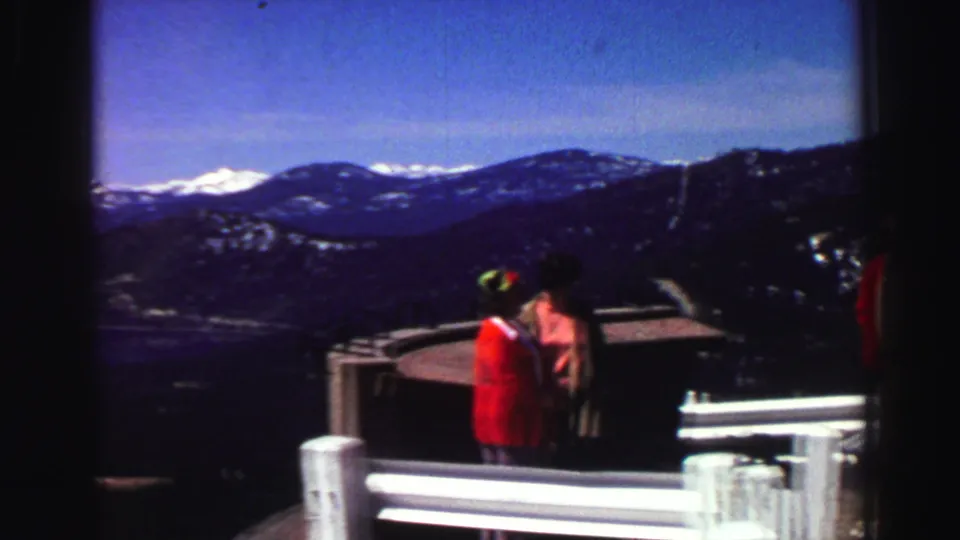 Two adults at a Lake Tahoe roadside viewpoint face the mountain landscape, with white guardrails, dark slopes, and snowy peaks visible.