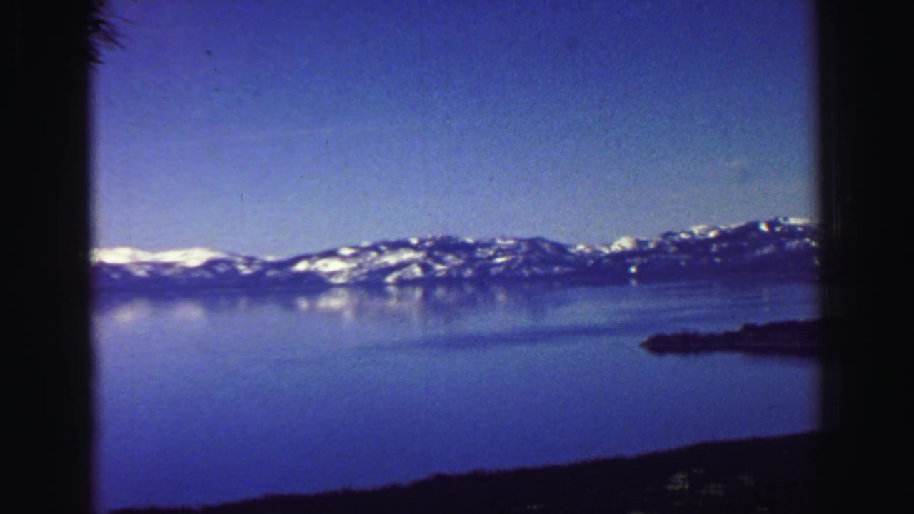 Lake Tahoe with snow-capped mountains across calm blue water, framed by dark pine silhouettes.