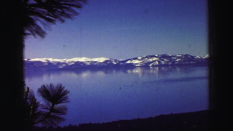 Lake Tahoe landscape in 1969 home-movie footage, with snow-capped mountains, blue water, and foreground pine silhouettes.