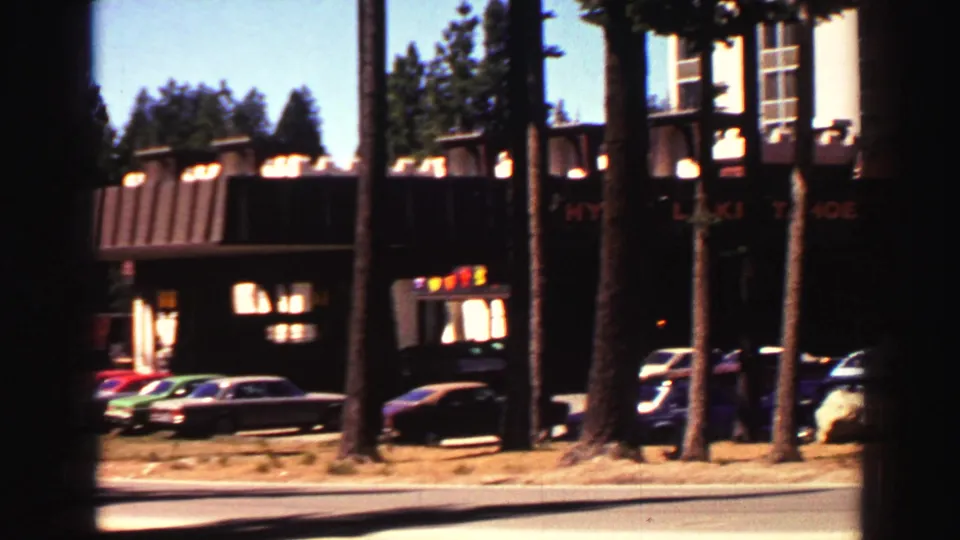 1969 Lake Tahoe hotel frontage with classic cars, roadside pines, dark awnings, and partially visible restaurant lettering.