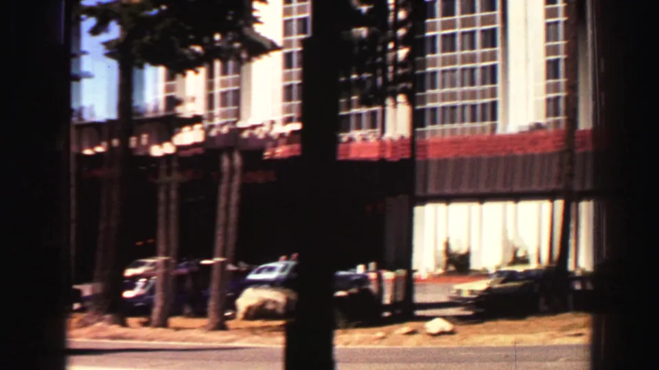 Lake Tahoe hotel exterior showing tall windows, red balcony railings, parked cars, and pine trunks near the road.