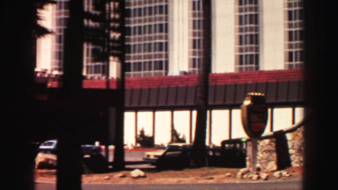 Hotel exterior with tall windows, red roofline, parked cars, pine trunks, and a shadowed foreground.