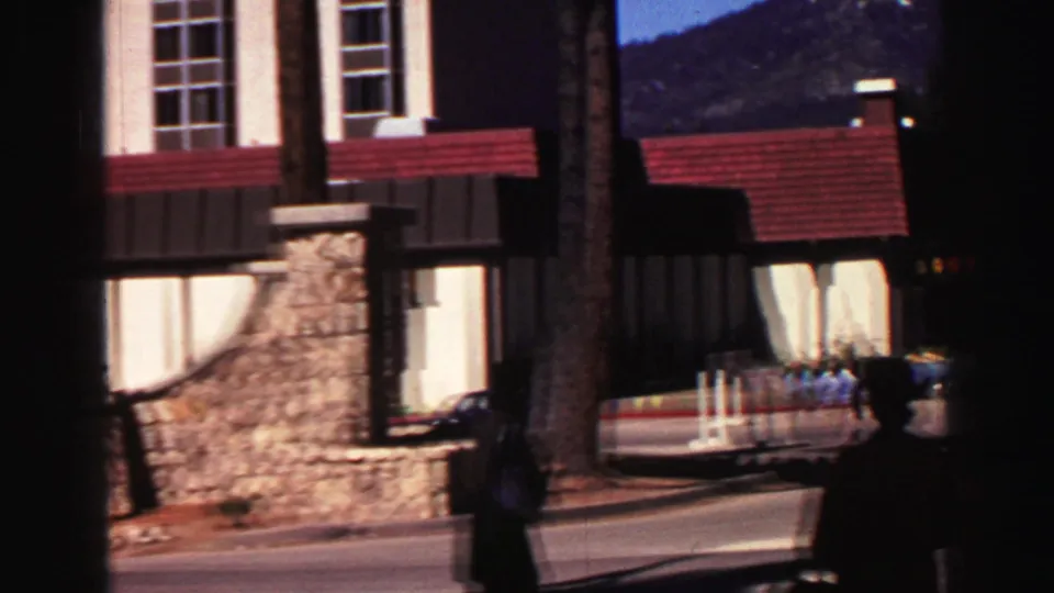 Lake Tahoe hotel corner and roadside area with a stone base, red roofline, pine trunks, and a shadowed person in 1969 home-movie footage.