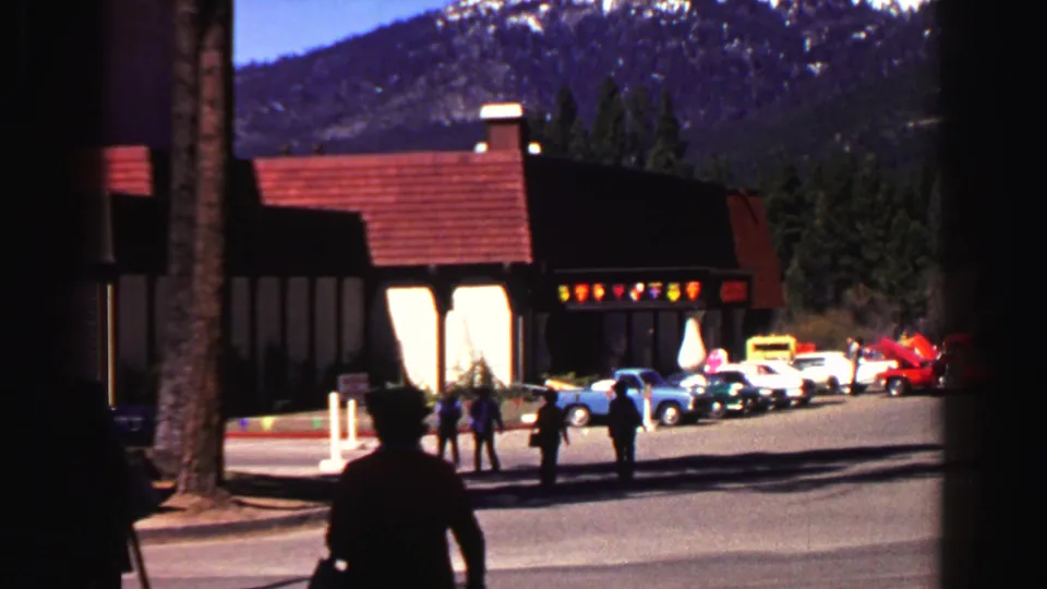 1969 Lake Tahoe parking lot with pedestrians, parked cars, resort buildings, pine trees, and snow-dusted mountains in the background.