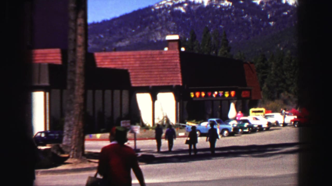 Adults approach a lodge entrance with parked cars and snowy mountains behind it.