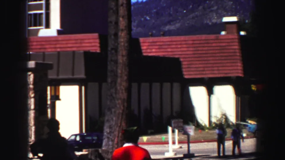 Adults gather near a Lake Tahoe lodge exterior in 1969, with period cars, trees, signage, and mountain slopes in view.