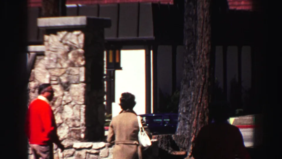 1969 Lake Tahoe frame showing two adults in period clothing walking near a stone-faced lodge and parked car.