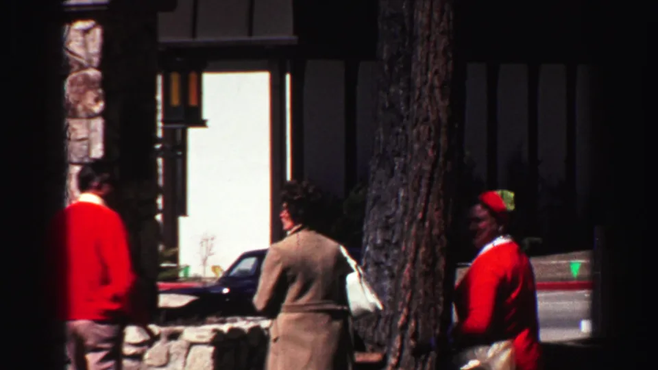 Adults meet outside a Lake Tahoe lodge in 1969, framed by trees, a stone wall, a parked car, and dark lodge siding.