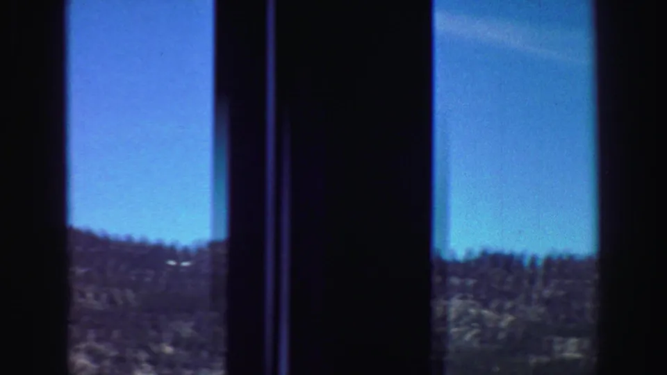 Final 1969 road-trip frame with dark window mullions, bright blue sky, distant hills, and a small trace of vegetation below.