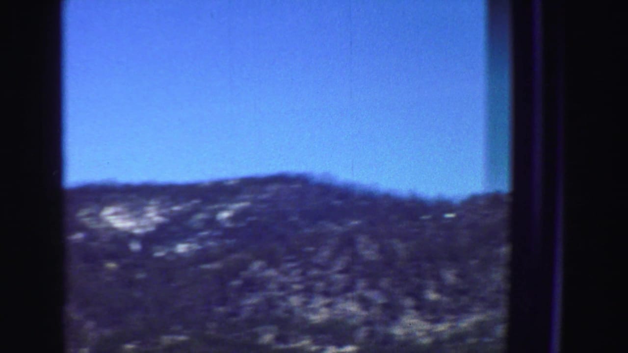 Forested mountain ridge seen through a dark window or film-frame border under blue sky.