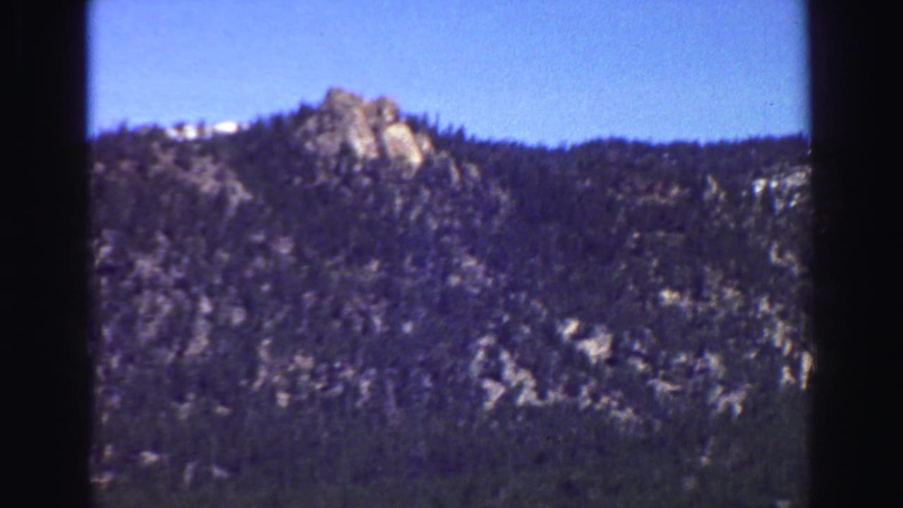 Rocky forested mountain slope under clear blue sky with dark vertical frame edges.