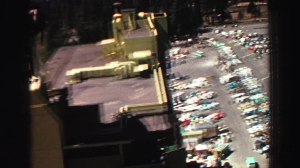 Elevated Lake Tahoe frame from 1969 showing a large building, access road, and rows of late-1960s cars in a busy lot.