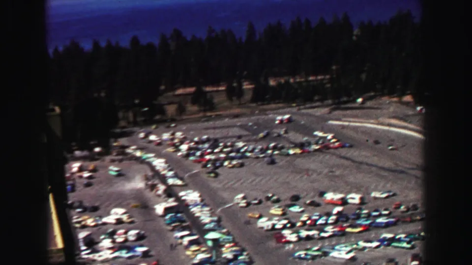 Wide 1969 Lake Tahoe view over packed parking areas, tree lines, hills, and small distant vehicles under a blue sky.
