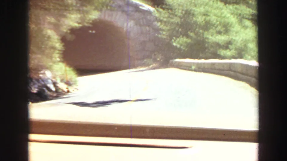 Lake Tahoe-area 1969 road footage showing a sunlit paved road leading toward a stone tunnel entrance and roadside wall.