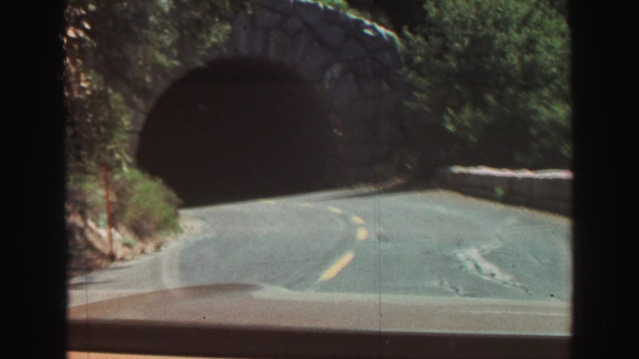 Road curves toward a dark stone tunnel entrance seen through a windshield.