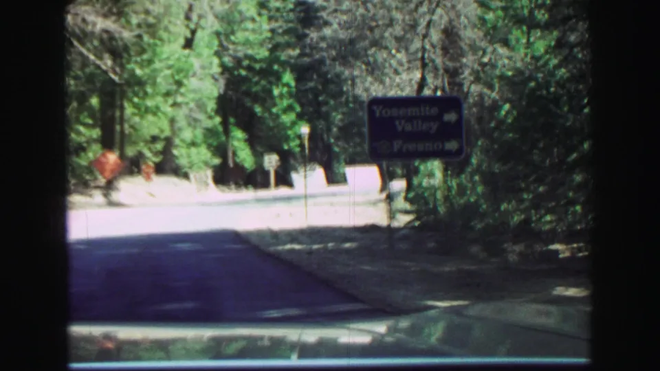 8mm frame from a 1969 California road trip showing a blue directional sign for Yosemite Valley and Fresno beside a wooded road.