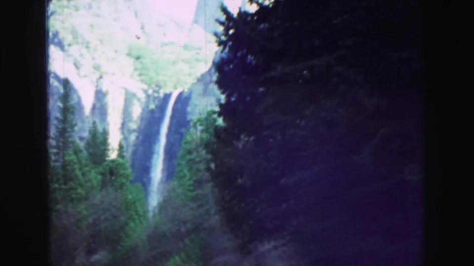 1969 8mm travel view of a tall waterfall beyond evergreens, seen from a moving vehicle or roadside viewpoint.