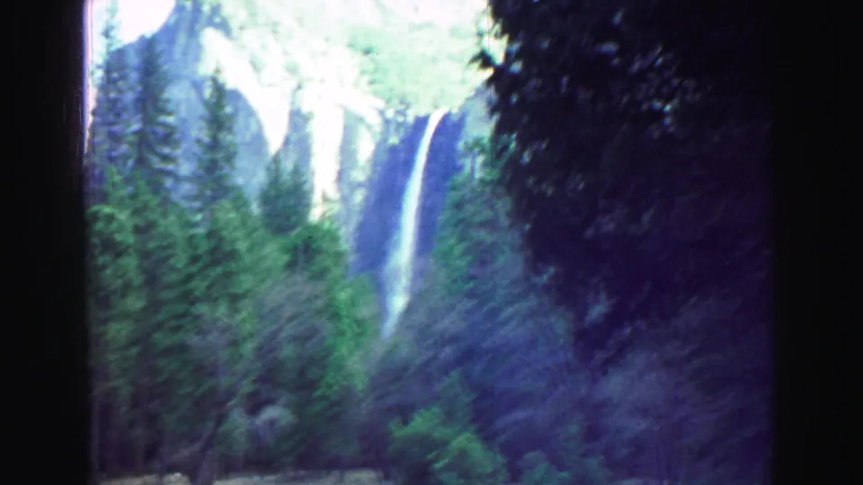 Vintage 8mm frame of a waterfall dropping down a rocky cliff, partially framed by tall trees and shadowed foreground foliage.