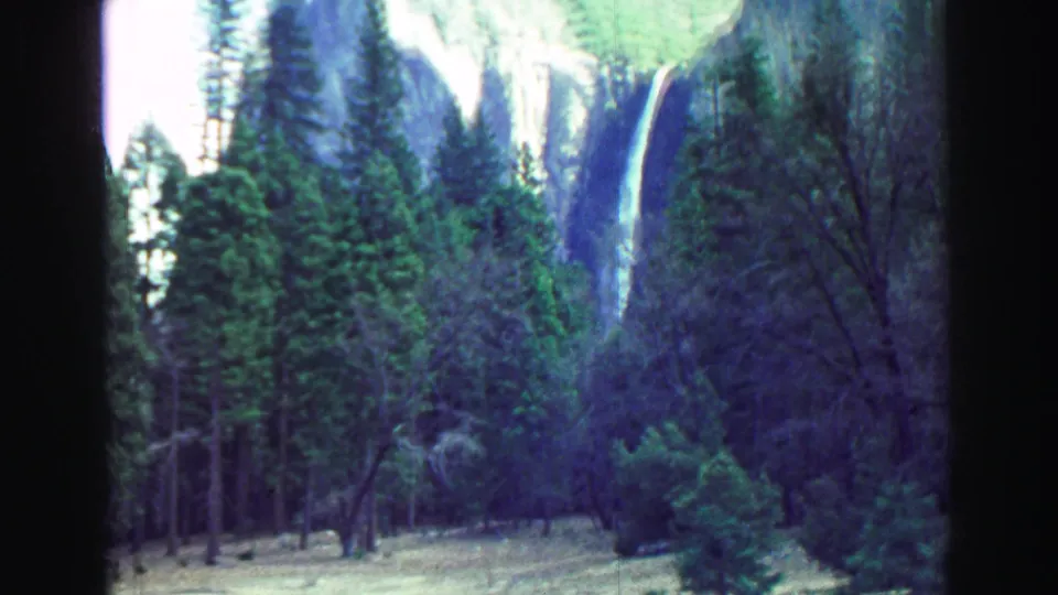 1969 8mm frame showing a waterfall behind dense evergreens, with dark film or vehicle framing at the sides.