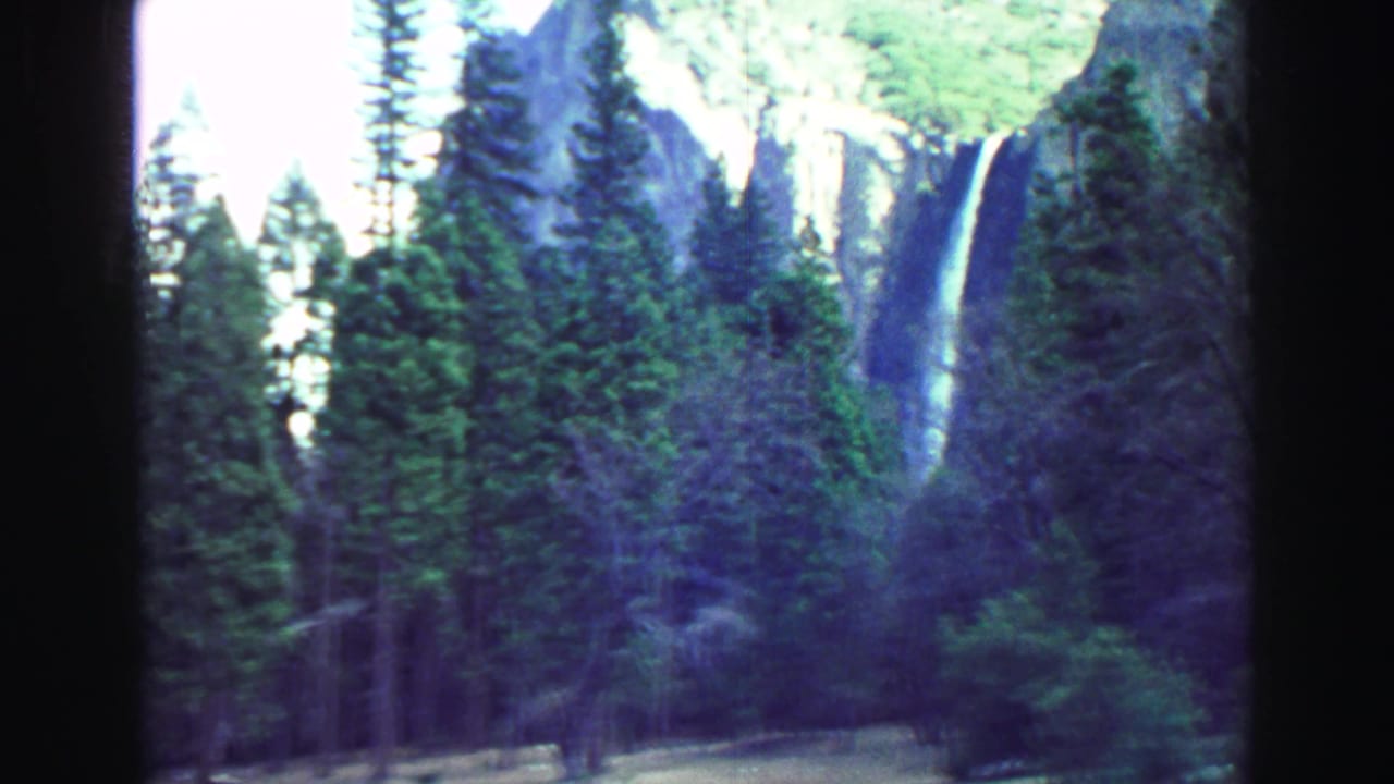 1969 8mm home-movie frame from the Lake Tahoe, California clip showing evergreen trees, a pale cliff, and a distant waterfall seen through the moving vehicle perspective.