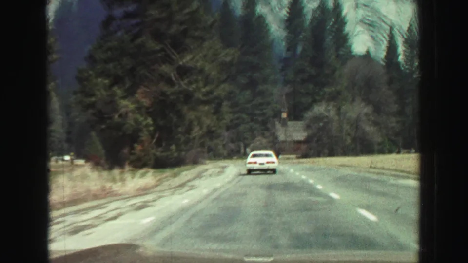 1969 Lake Tahoe-area road footage showing a white car ahead on a paved highway with evergreen trees, roadside grass, and mountain slopes in the background.