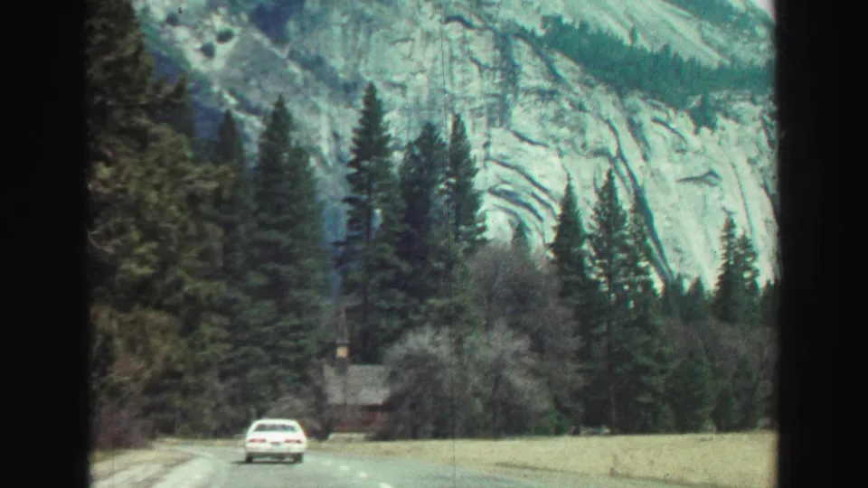 1969 8mm frame from a moving vehicle showing a white car on a California mountain road below pale rock faces, conifers, and a small roadside structure in the distance.