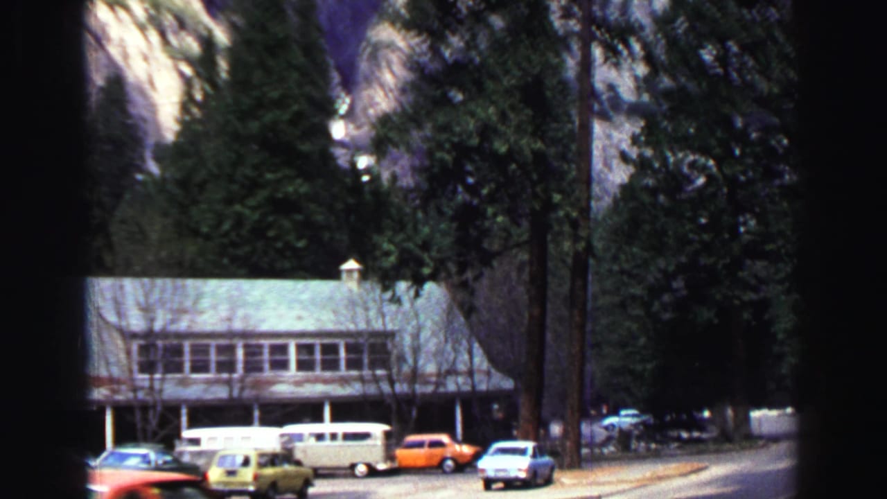 Rustic roadside lodge with parked cars beneath tall trees and mountain cliffs.