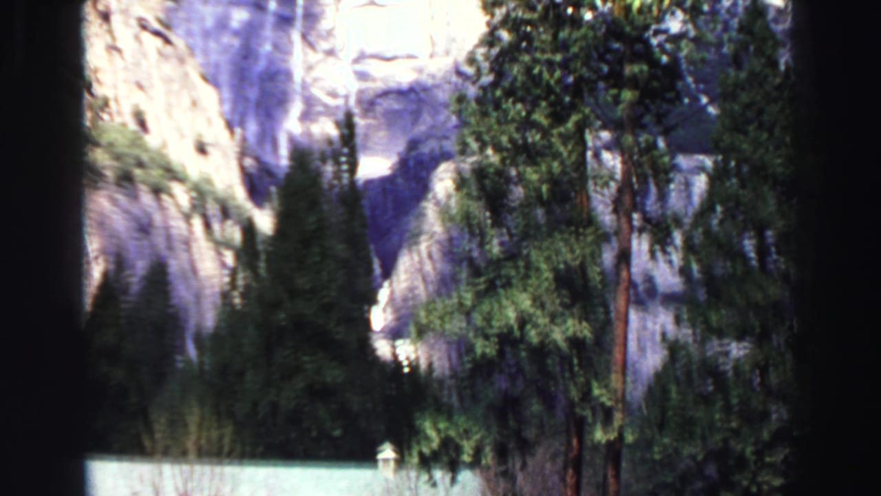Evergreen trees stand in front of sunlit granite cliffs and a narrow waterfall.