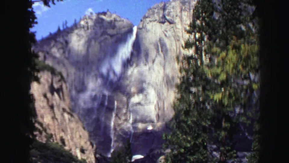 Lake Tahoe 1969 home-movie frame of a waterfall crossing a granite cliff, framed by blue sky and dark evergreens.